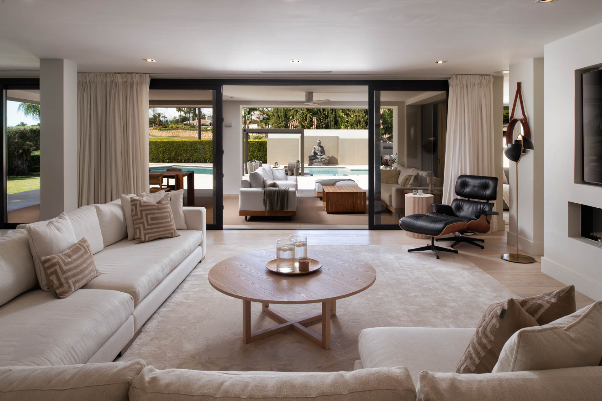 The living room with pale floors and soft neutral tones, open to the covered terrace and pool beyond.
